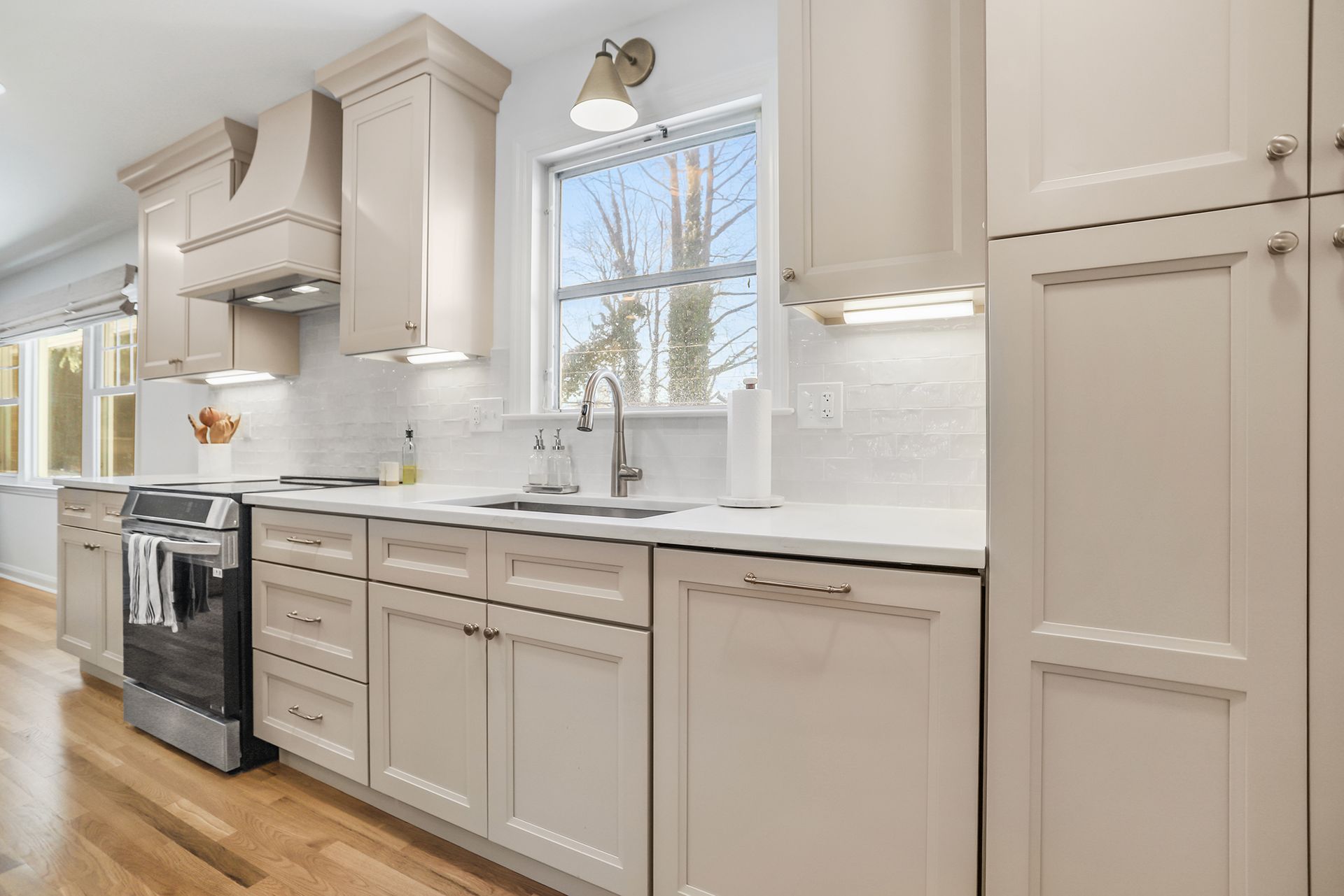 Bright white kitchen with beige cabinets, marble counters, stainless stove, and a window over the sink
