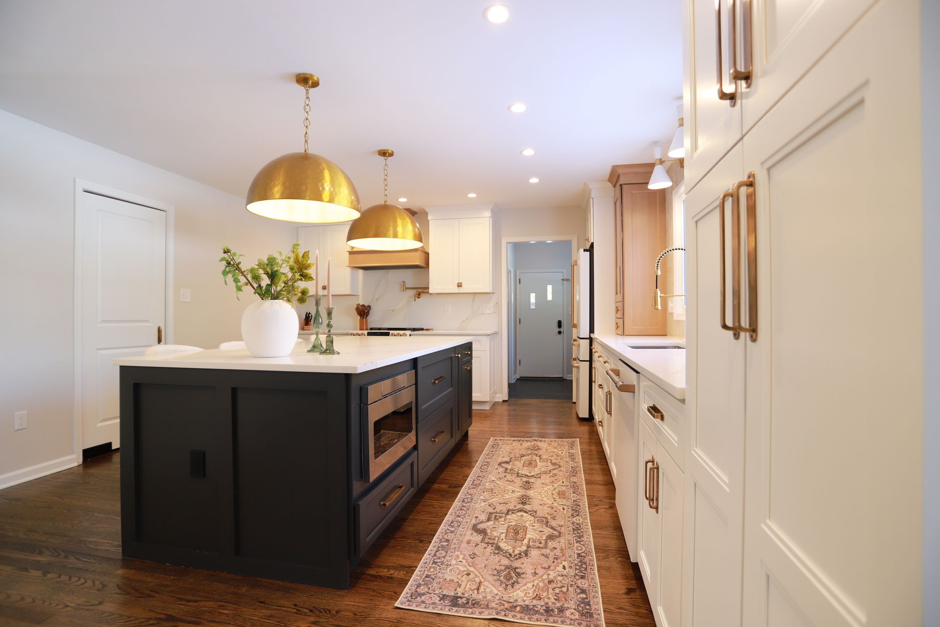 Modern kitchen with a dark island, brass pendant lights, white cabinetry, and a patterned runner rug.