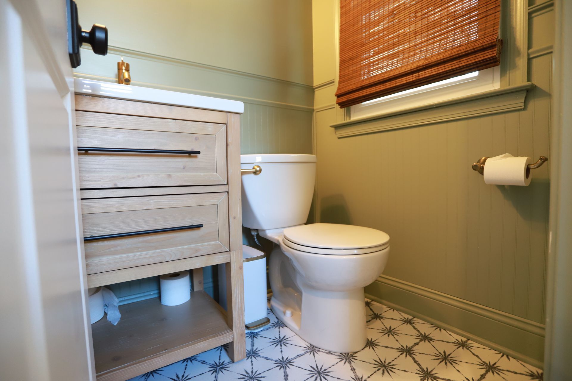 Small bathroom with toilet, white sink cabinet, patterned tile floor, and window with bamboo shade.