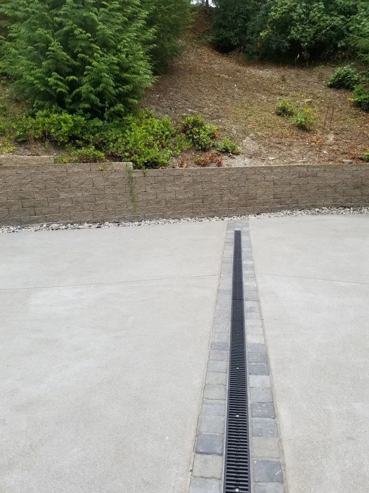 Concrete walkway with a narrow drainage grate leading to a landscaped slope with green shrubs.