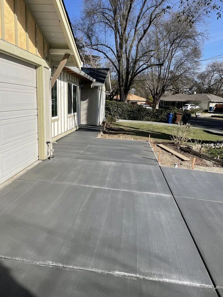 Concrete driveway beside a beige house, with a garage door, bare trees, and a yard in the background.