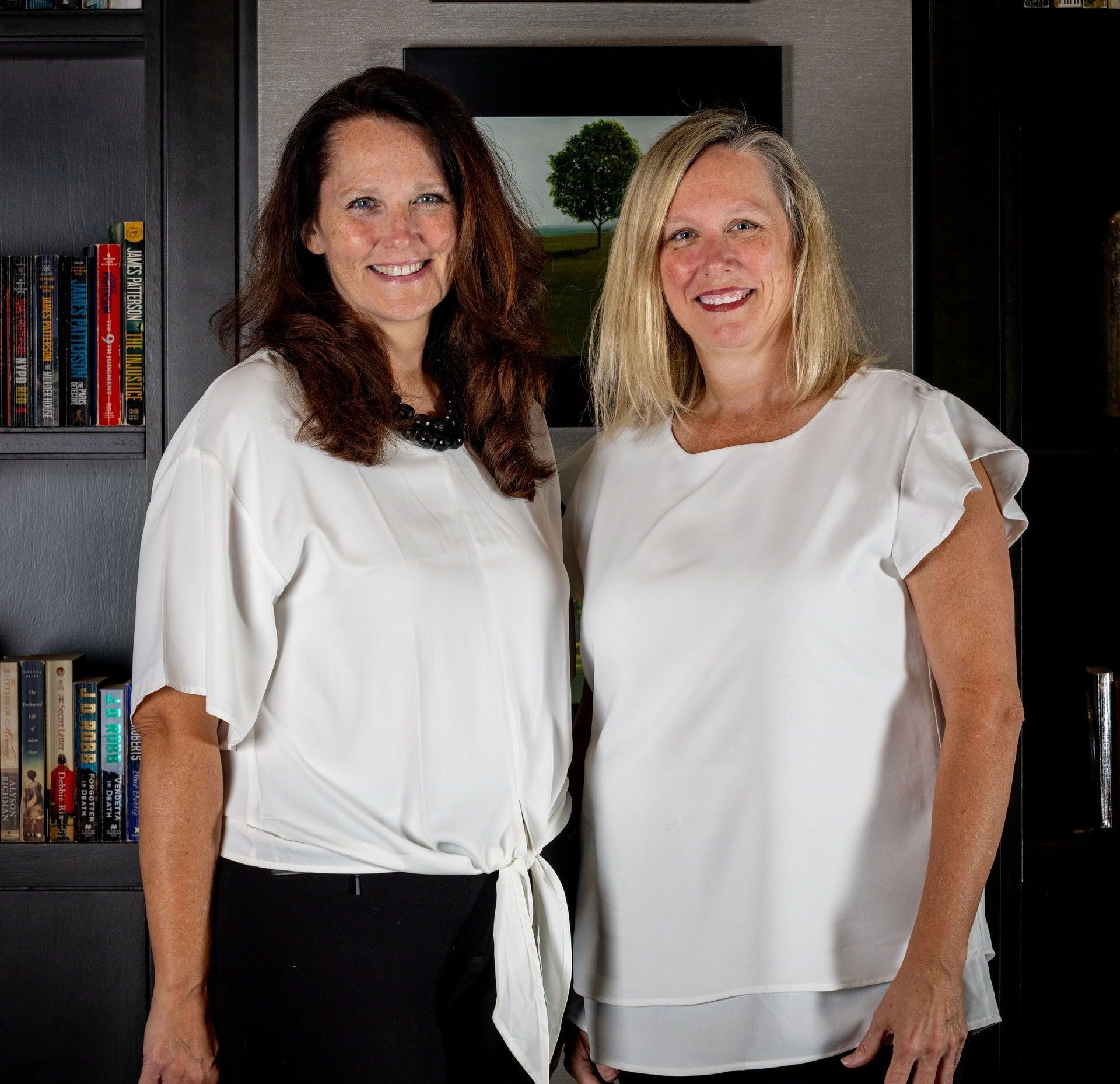 Two women standing in front of a bookshelf, smiling in white blouses.