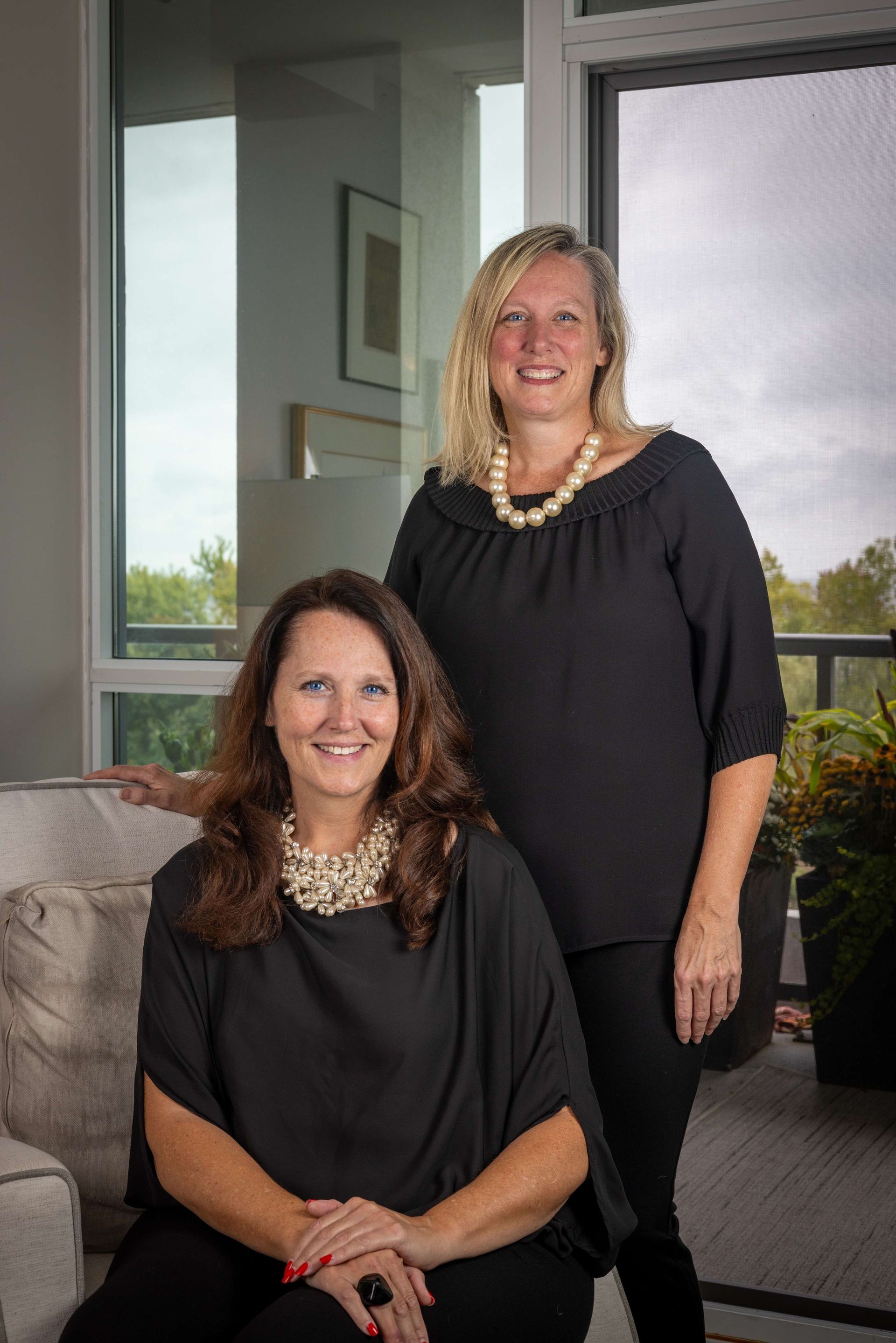 Two women posing in a bright office lounge, one seated and one standing, both in black outfits and necklaces