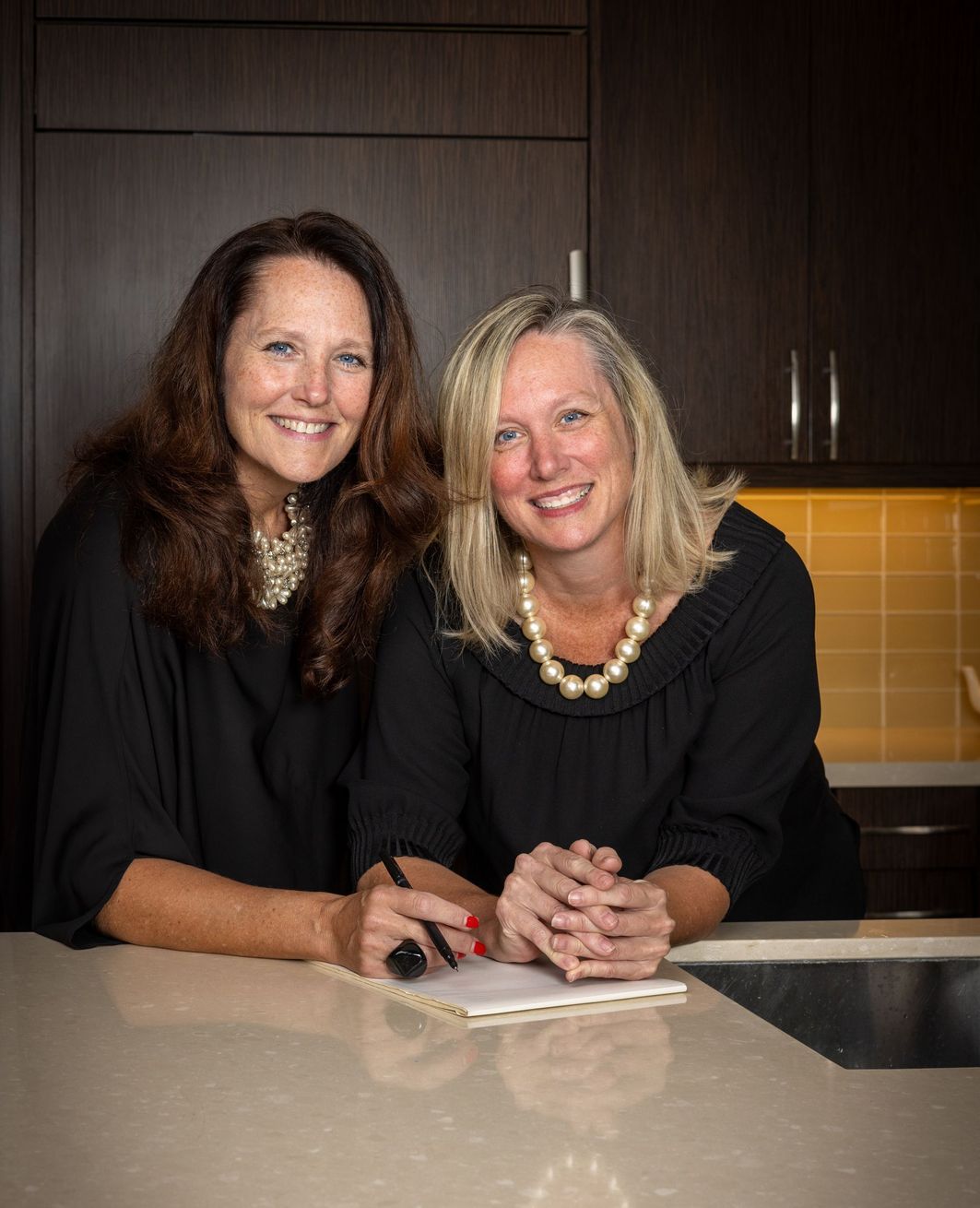 Two women smiling and posing in a kitchen, leaning on a counter.