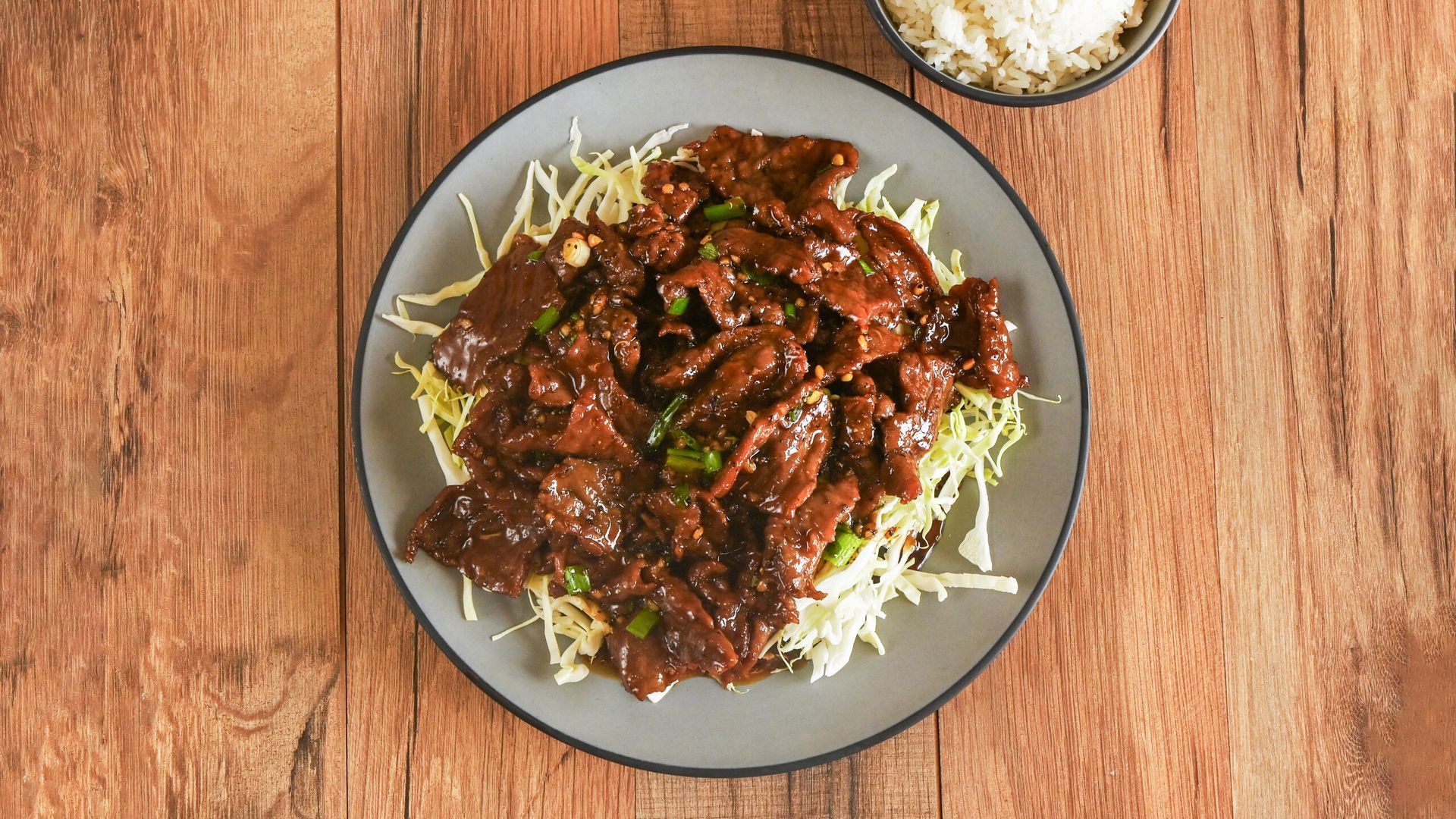 Plate of saucy meat over shredded cabbage on a wooden table, with a small bowl of rice nearby