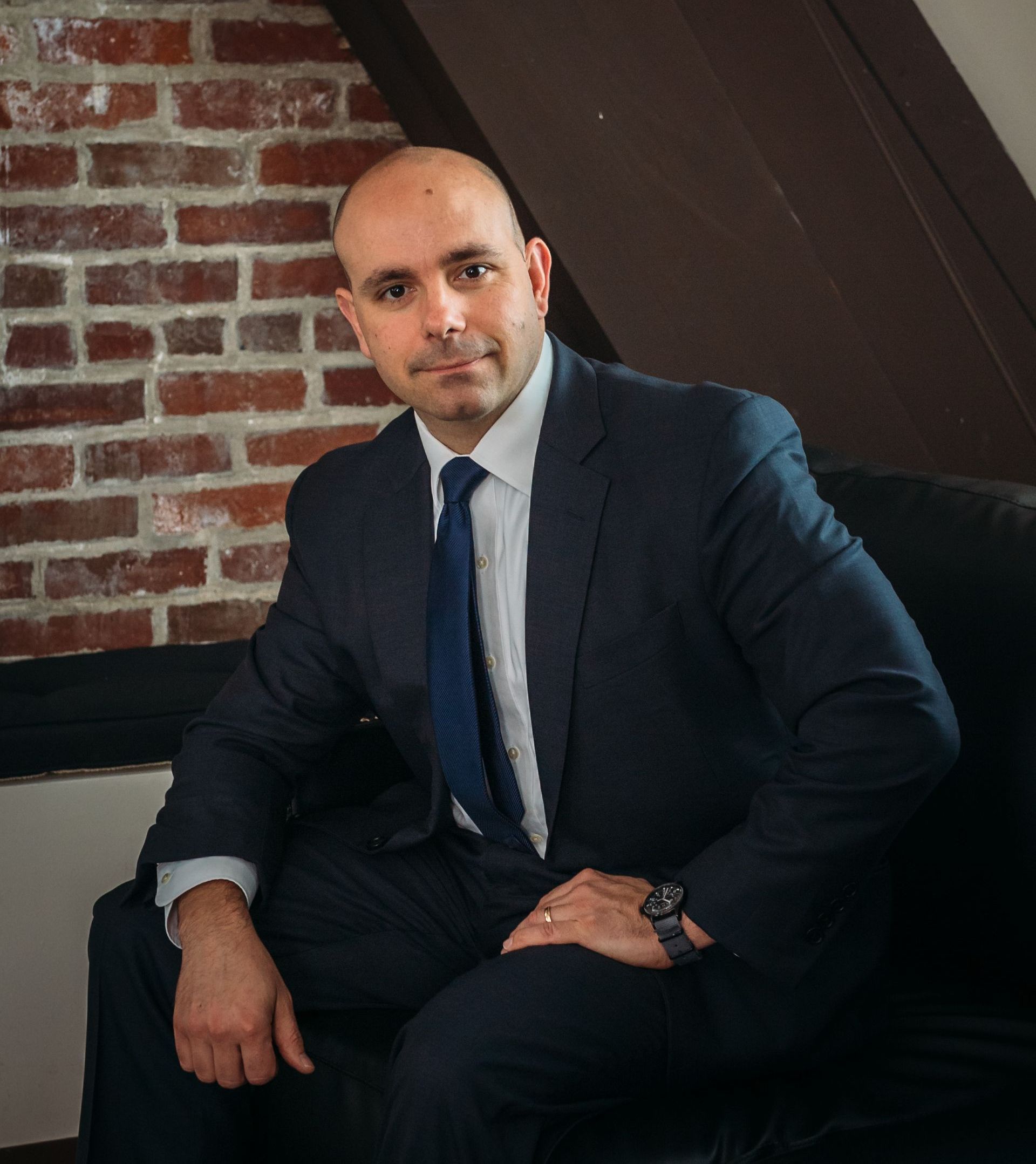 Man in suit sitting, looking at the camera. Brick wall background.