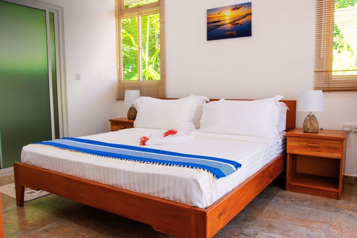 Bedroom with a wooden bed, nightstands, and a small painting on the wall. Natural light streams through the windows.