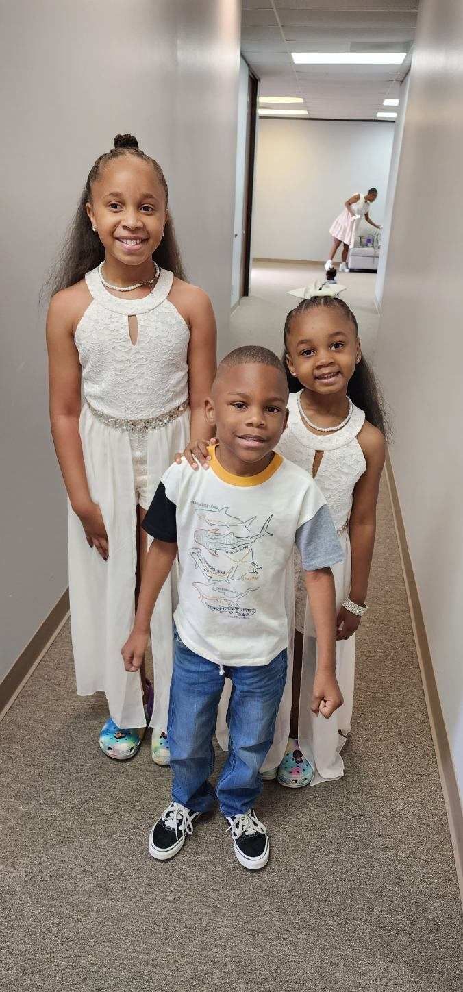 Three smiling children, two girls in white dresses, one boy in jeans and a t-shirt, in a hallway.