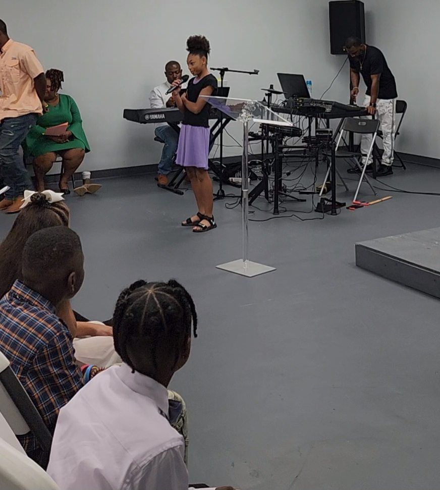 Girl speaking at a podium, possibly performing. Children seated, adults in the background with instruments.