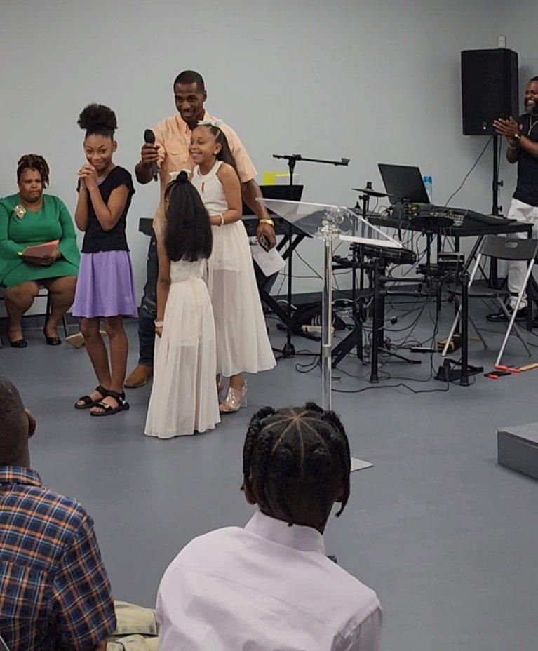 Man with mic and two girls in white dresses on a stage; audience watches.