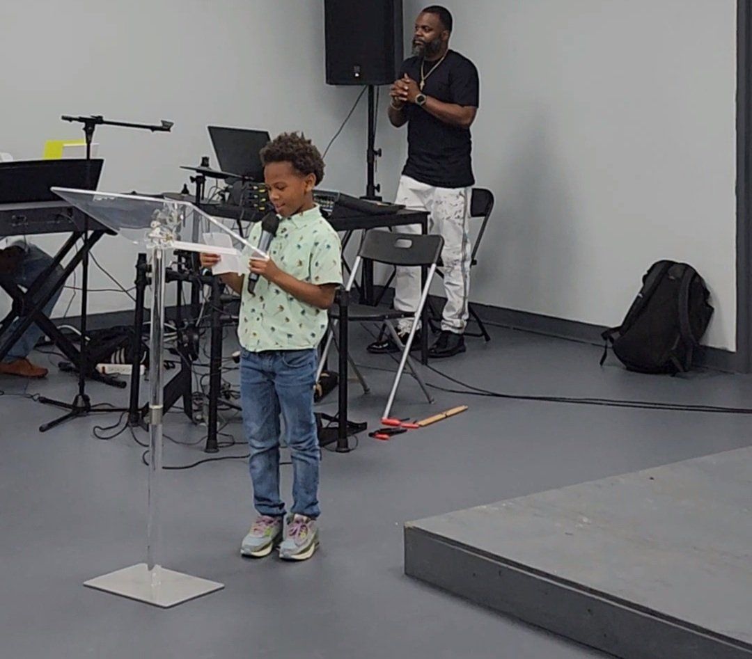 Young boy reading at a podium, man behind him with hands clasped, band equipment in background.