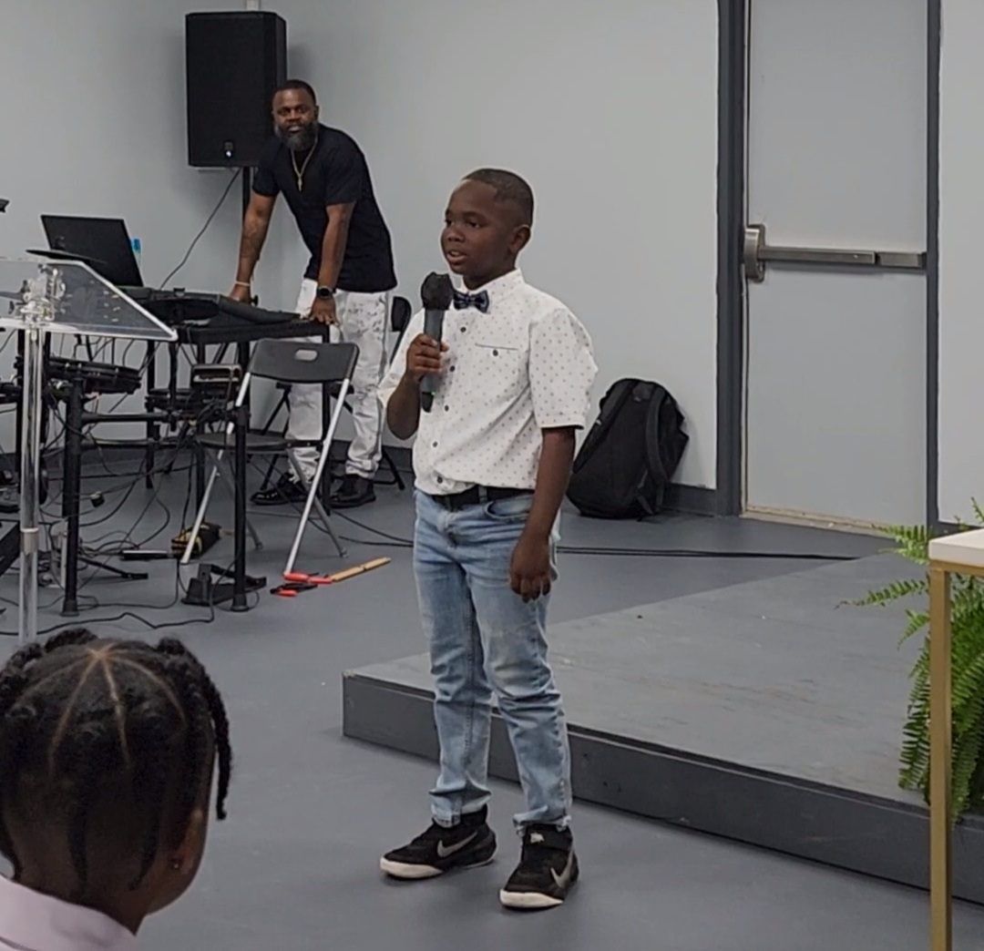 Young boy in bow tie speaks into microphone on stage; man at keyboard in background.