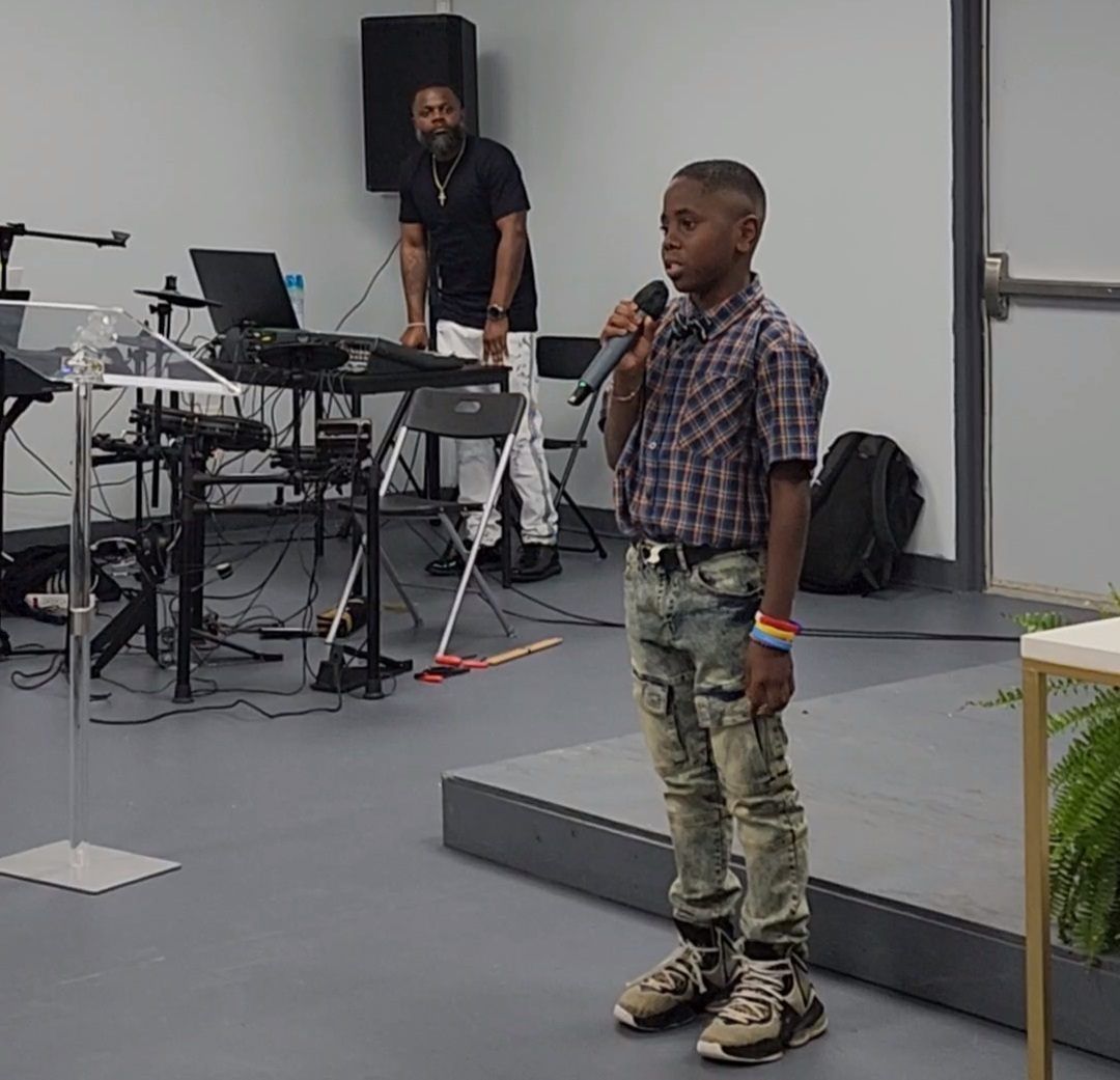 A young boy speaking into a microphone on a stage; a man is at a keyboard in the background.