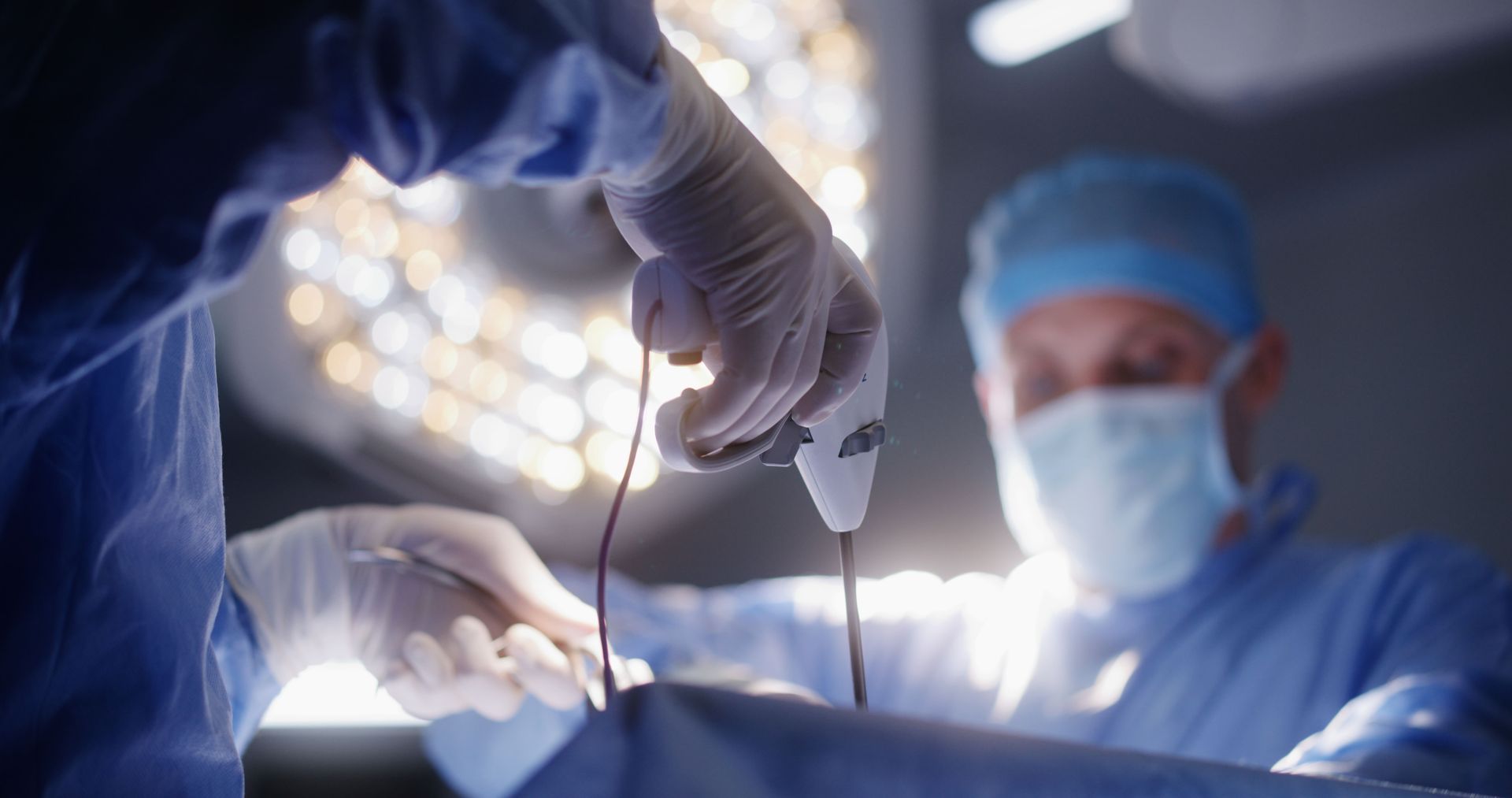 Cirurgiões de jaleco azul realizando uma operação sob luzes brilhantes de sala de cirurgia.