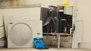 An air conditioning unit under repair with manifold gauges attached, a blue tool case, and a yellow hard hat nearby.