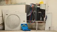An air conditioning unit under repair with manifold gauges attached, a blue tool case, and a yellow hard hat nearby.