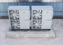 Two gray industrial air conditioning units side-by-side on a concrete slab against a blue wall.