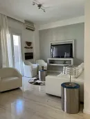 A modern living room with white armchairs, a white sofa, a metal side table with a lamp, and a wall-mounted television.