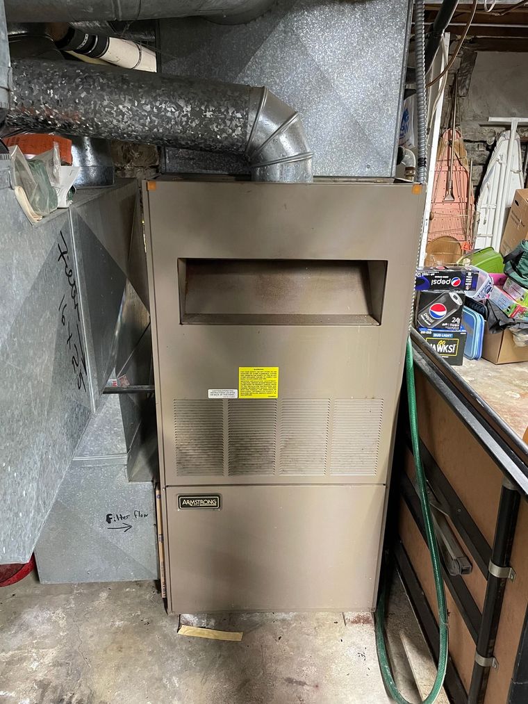 A tan residential furnace stands in a basement next to metal ductwork and a green hose.