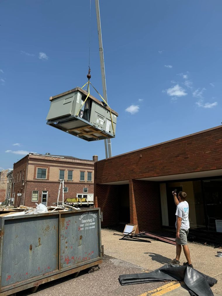 A crane lifts a large industrial HVAC unit onto a building roof as a worker in a white shirt directs the process.