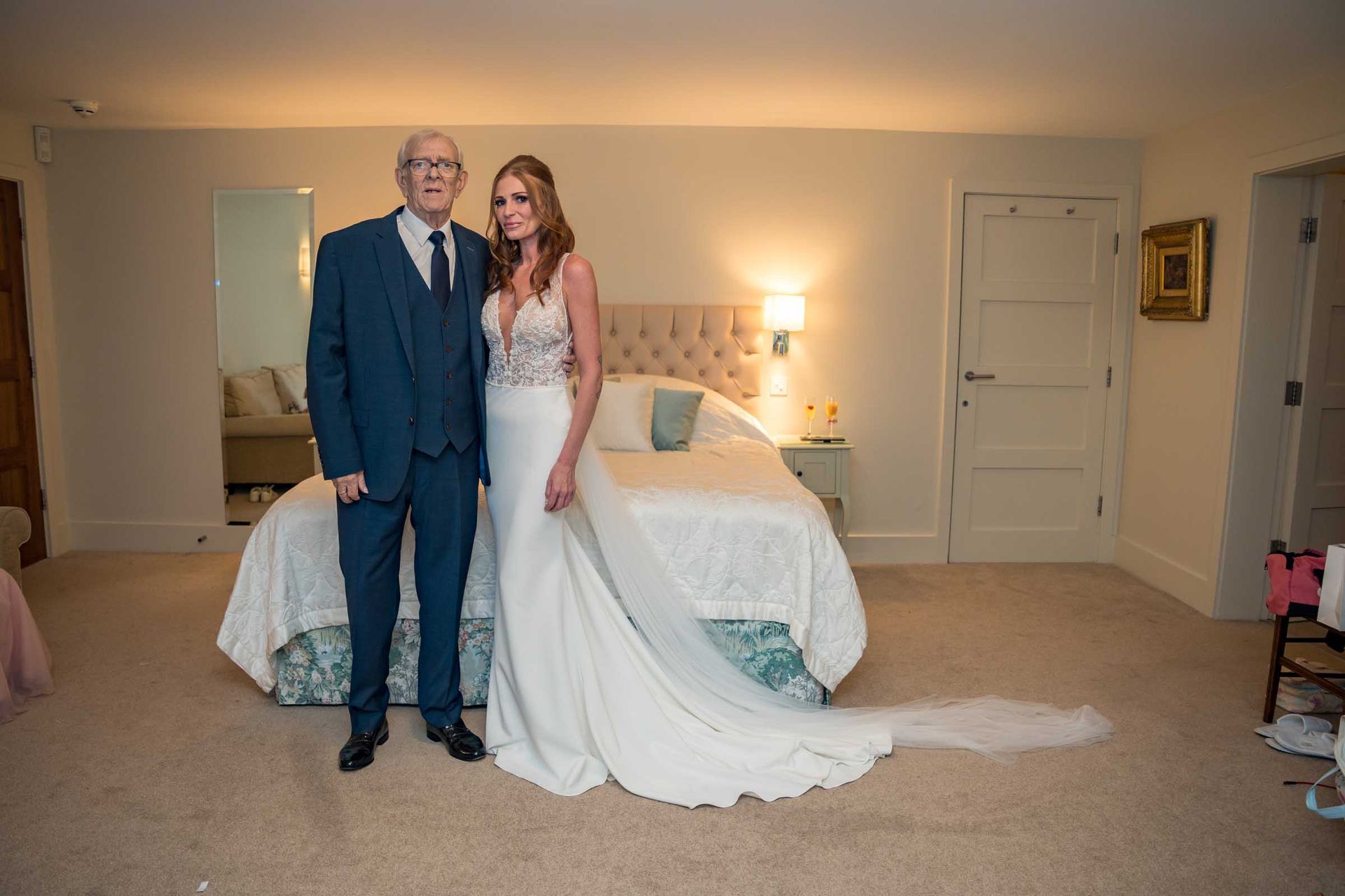 A bride and her father are posing for a picture in a bedroom.