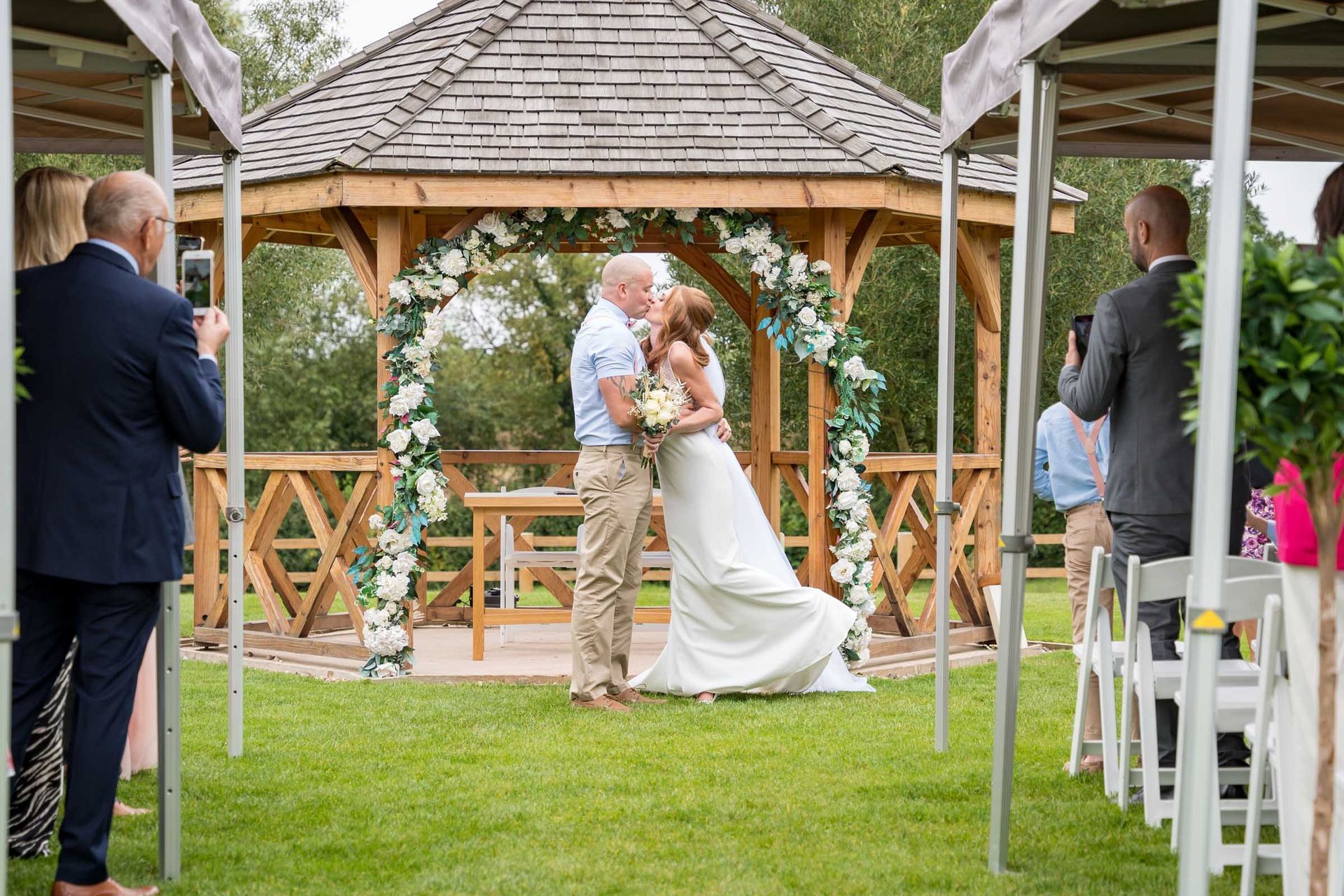 A man is taking a picture of a bride and groom kissing in front of a gazebo.