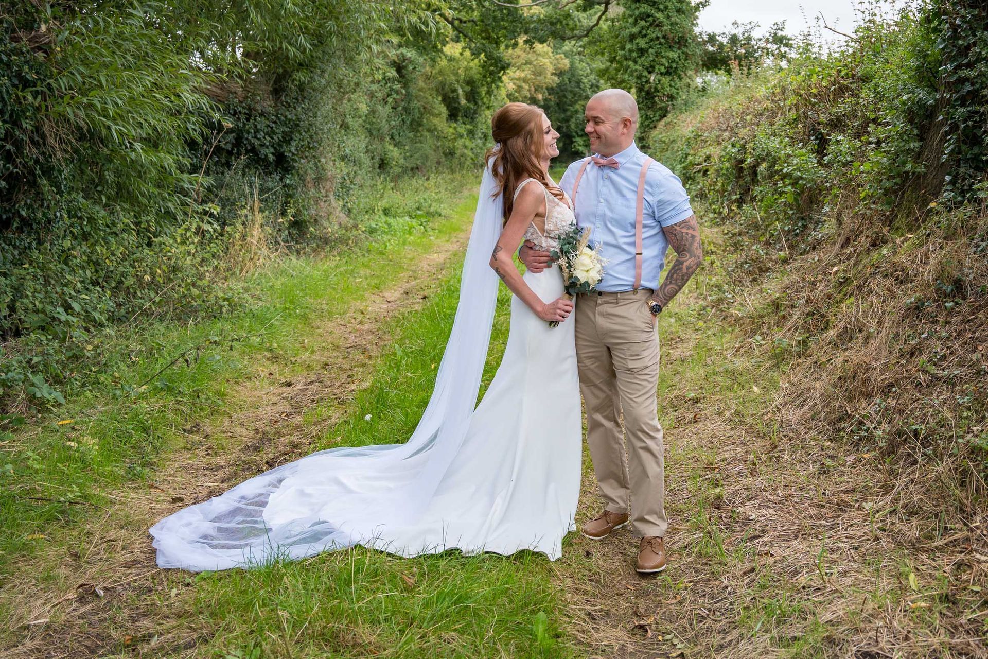 A bride and groom are standing next to each other on a path in the woods.