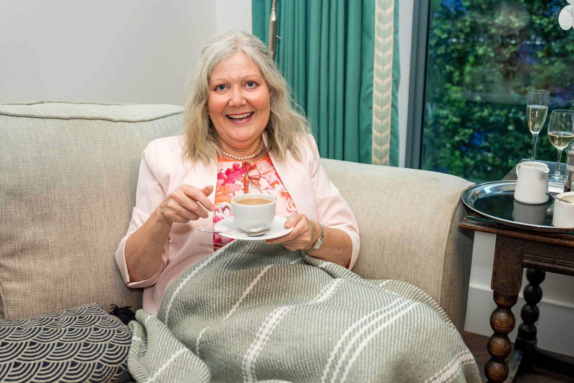 A woman is sitting on a couch covered in a blanket holding a cup of coffee.