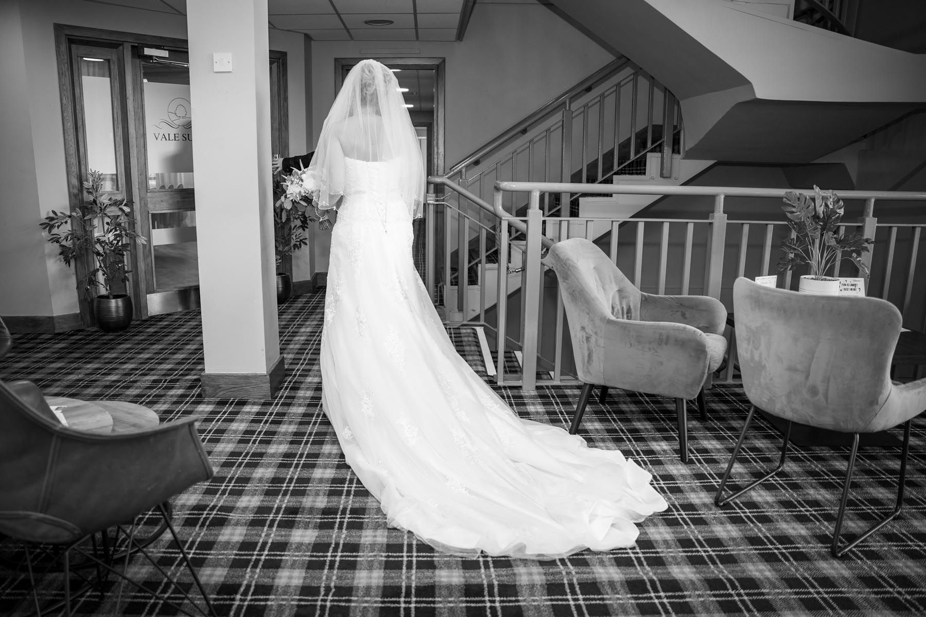 A black and white photo of a bride in a wedding dress standing in a hallway.