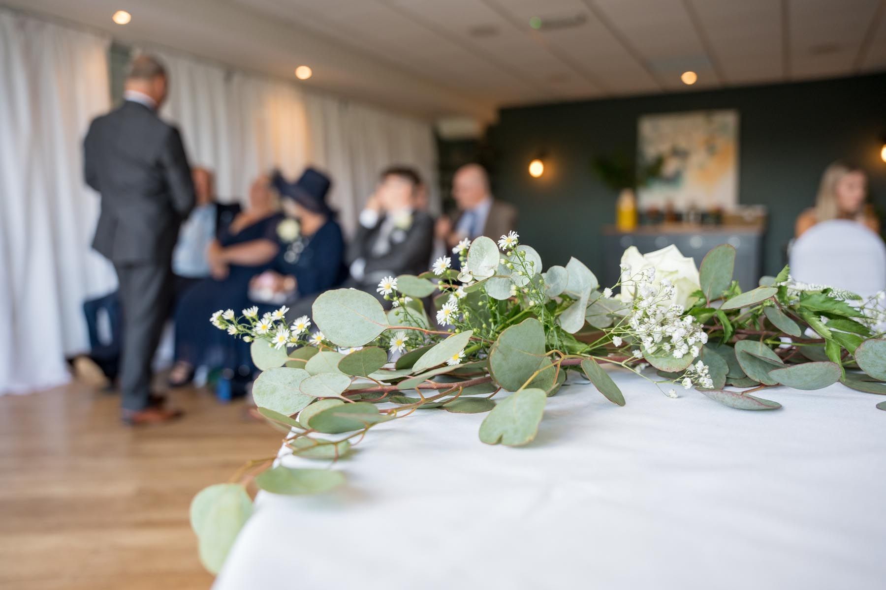 A table with flowers and leaves on it in a room with people in the background.