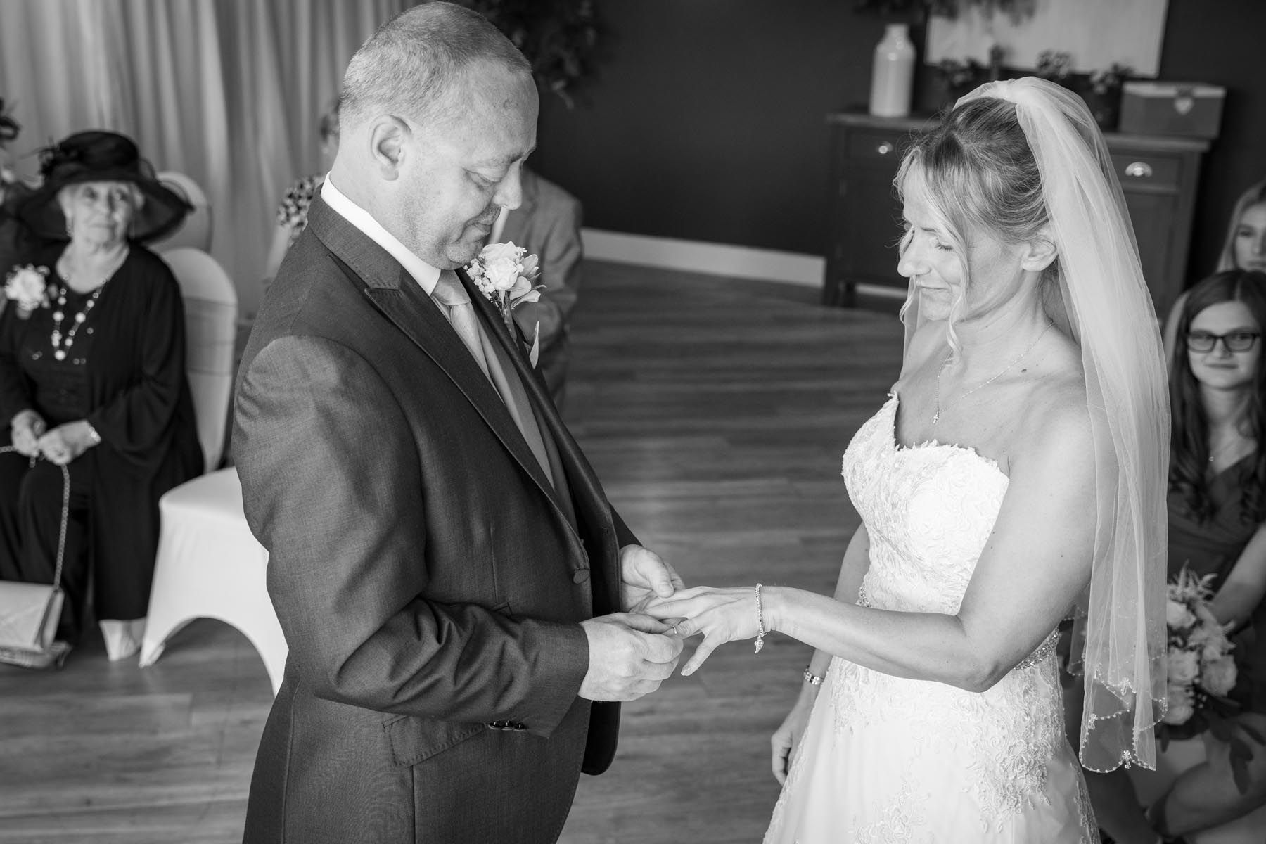 A black and white photo of a bride putting a wedding ring on her groom 's finger.