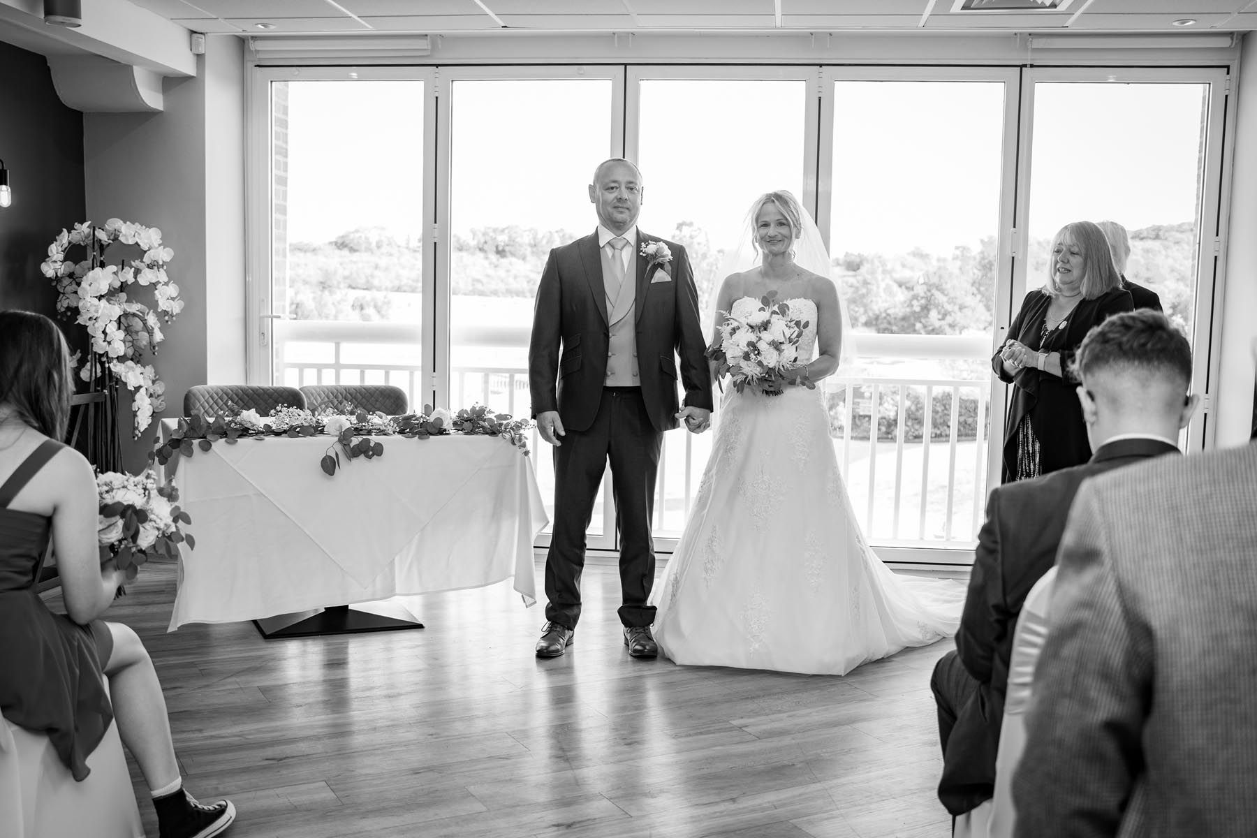 A black and white photo of a bride and groom holding hands during their wedding ceremony.