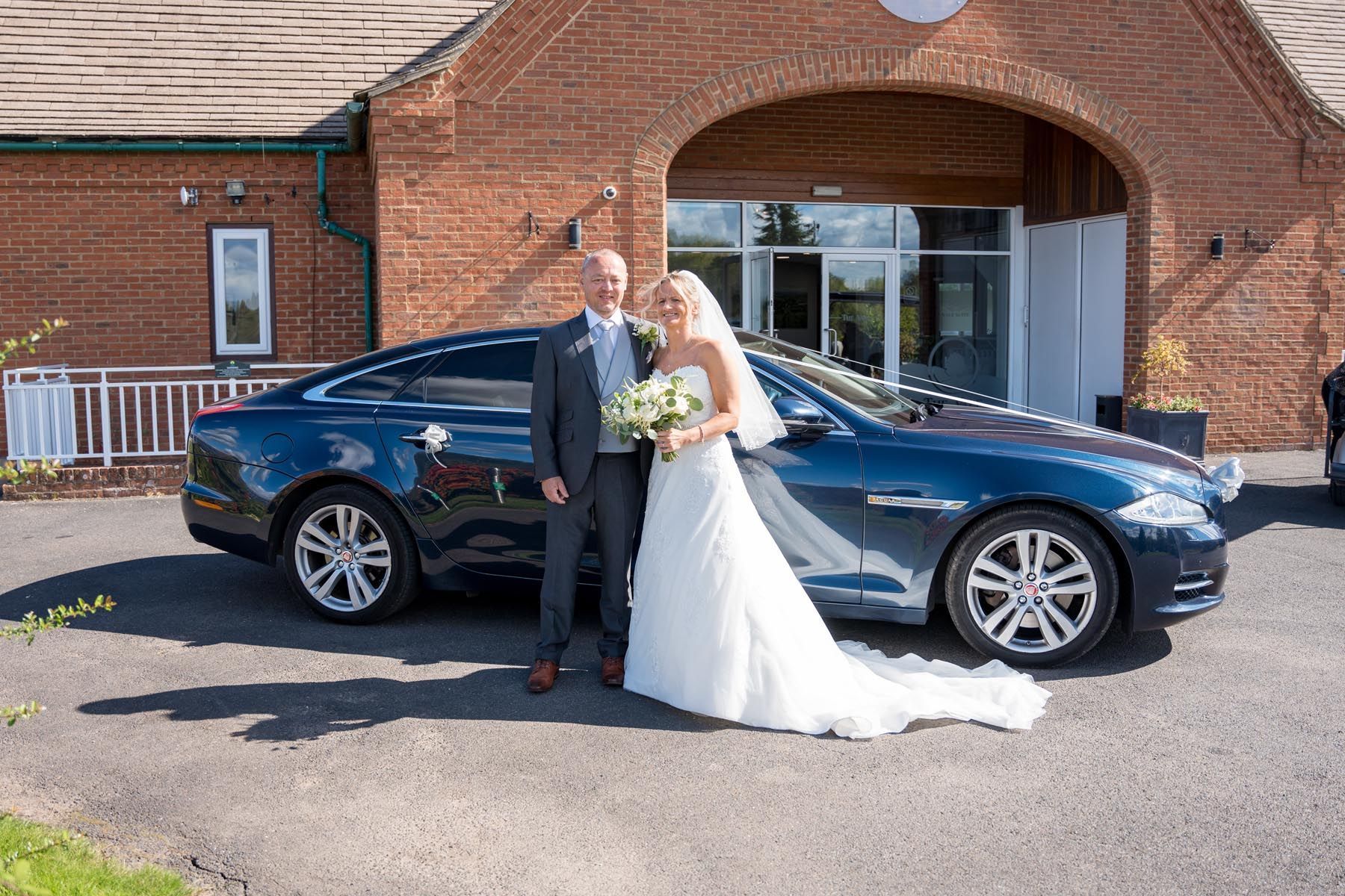 A bride and groom are posing for a picture in front of a car.