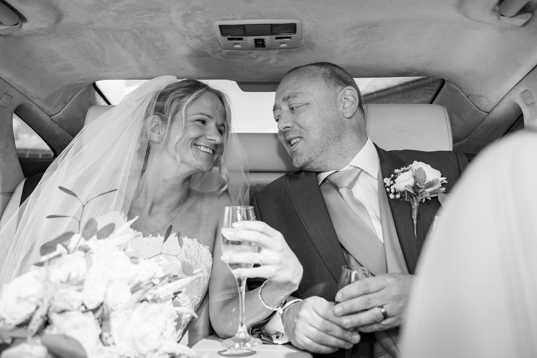 A bride and groom are sitting in the back seat of a car drinking wine.