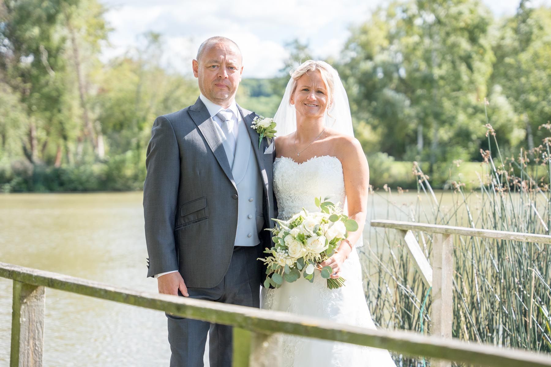 A bride and groom are posing for a picture on a bridge overlooking a lake.