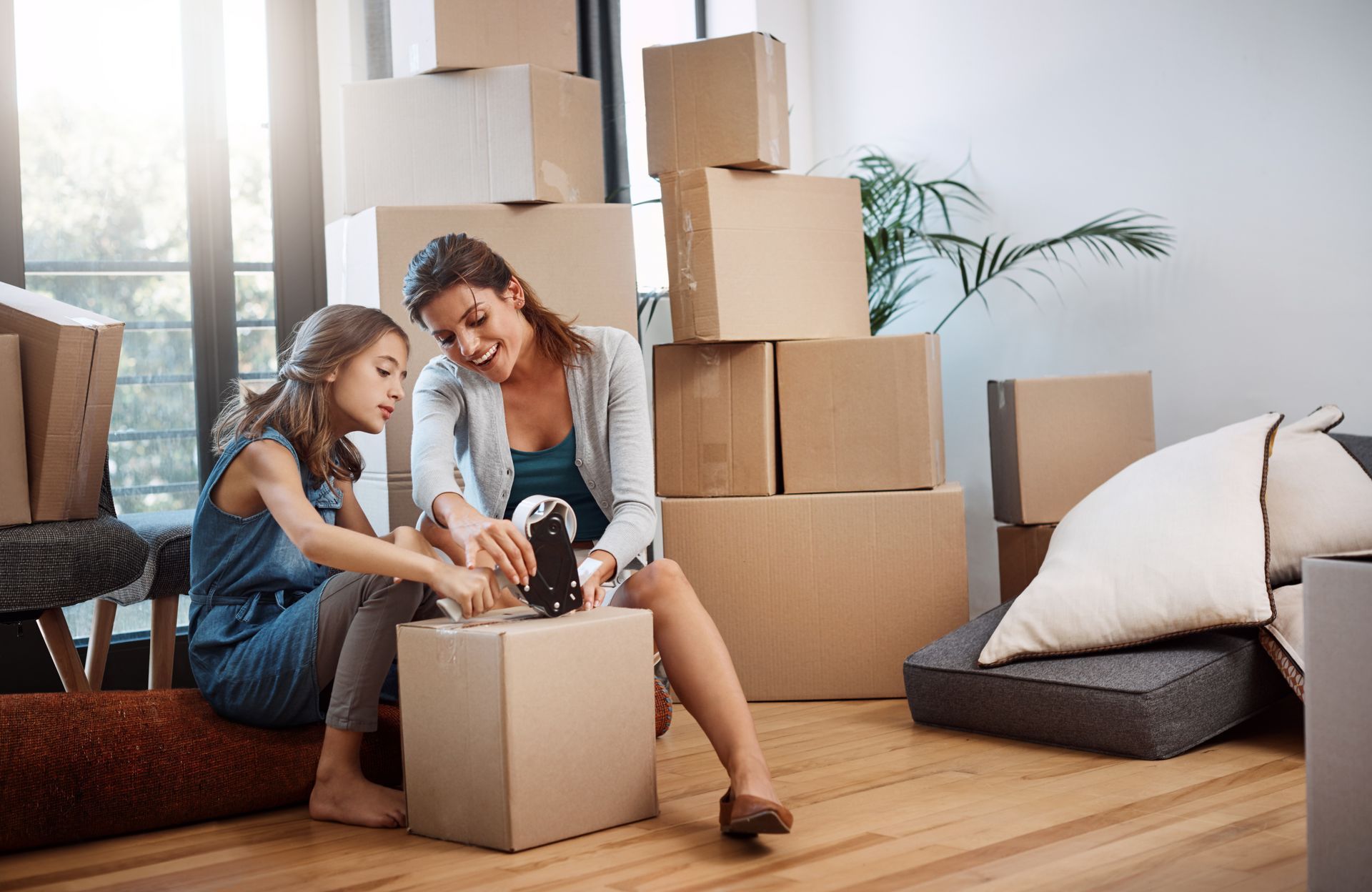 Mother and daughter smiling while taping a cardboard moving box together in a bright room.