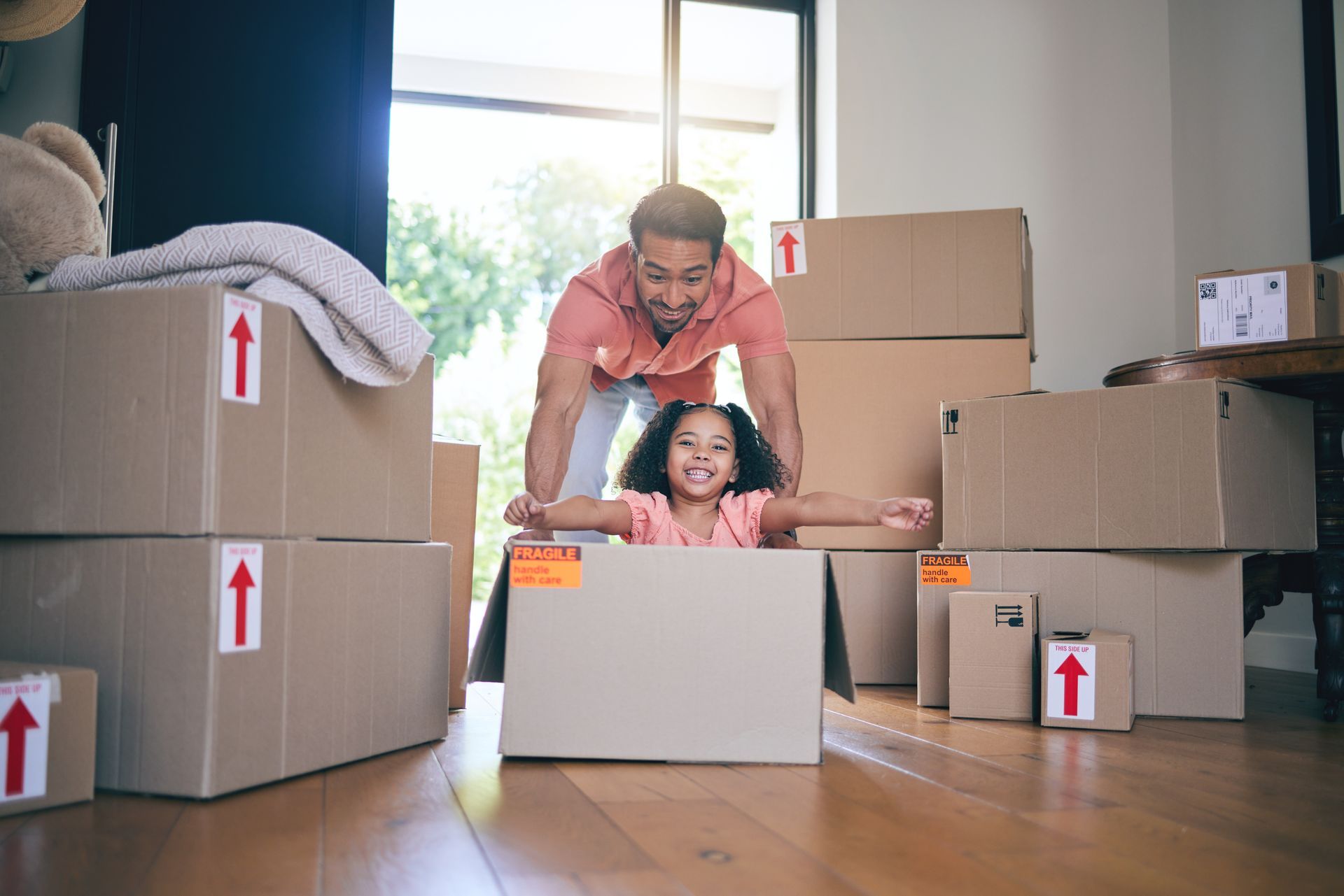 Smiling father playfully pushing his laughing daughter inside a cardboard box during a home move.
