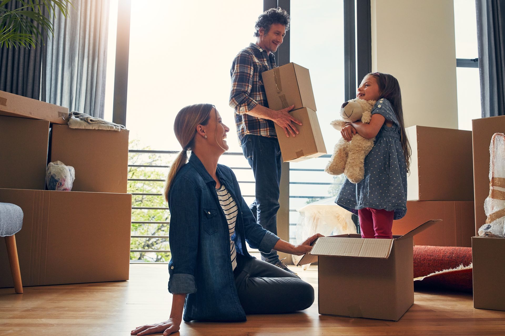 Family unpacking boxes in a new home; girl in box smiles, parents nearby.