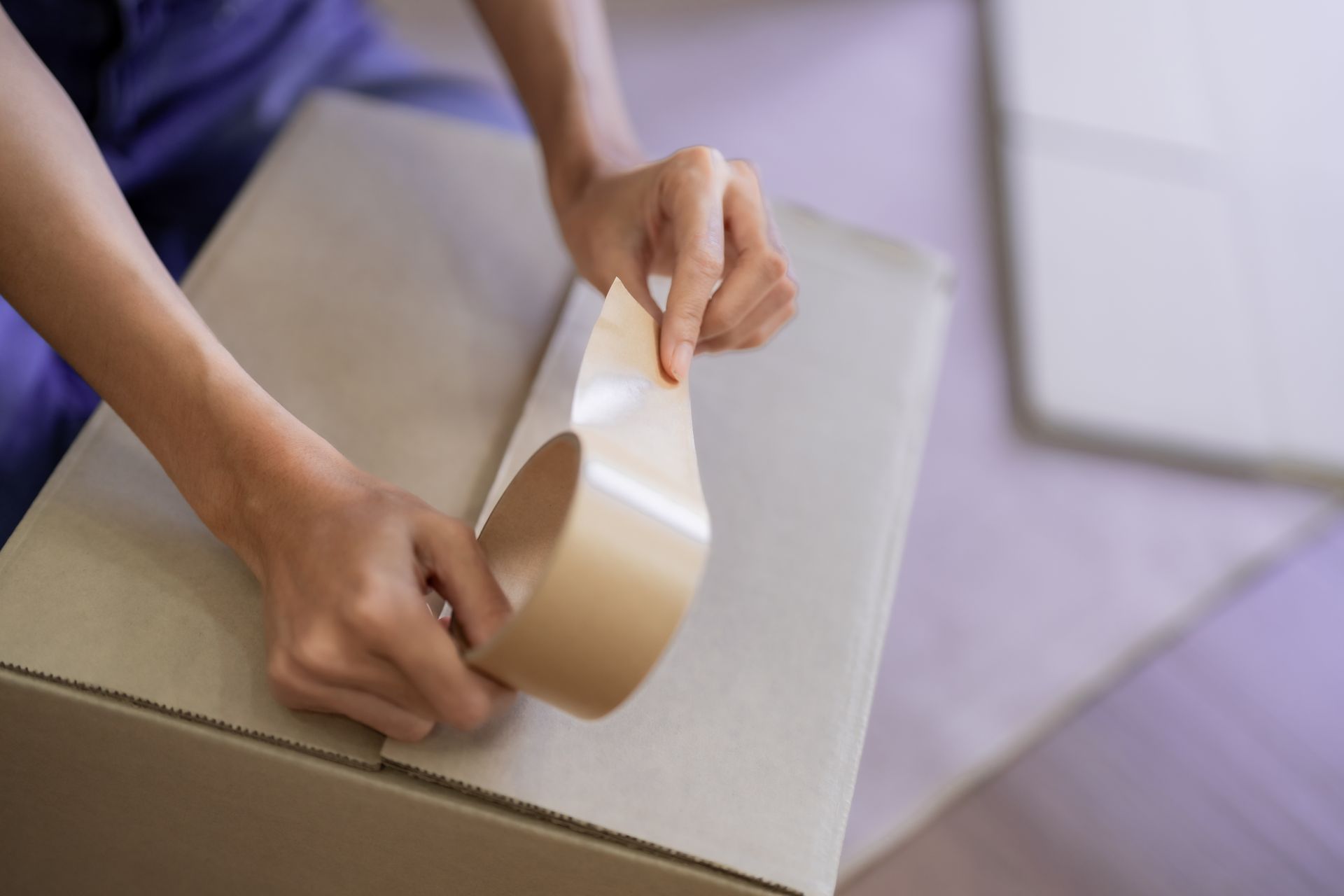 Close-up of a person's hands using a packing tape dispenser to seal a brown cardboard moving box.