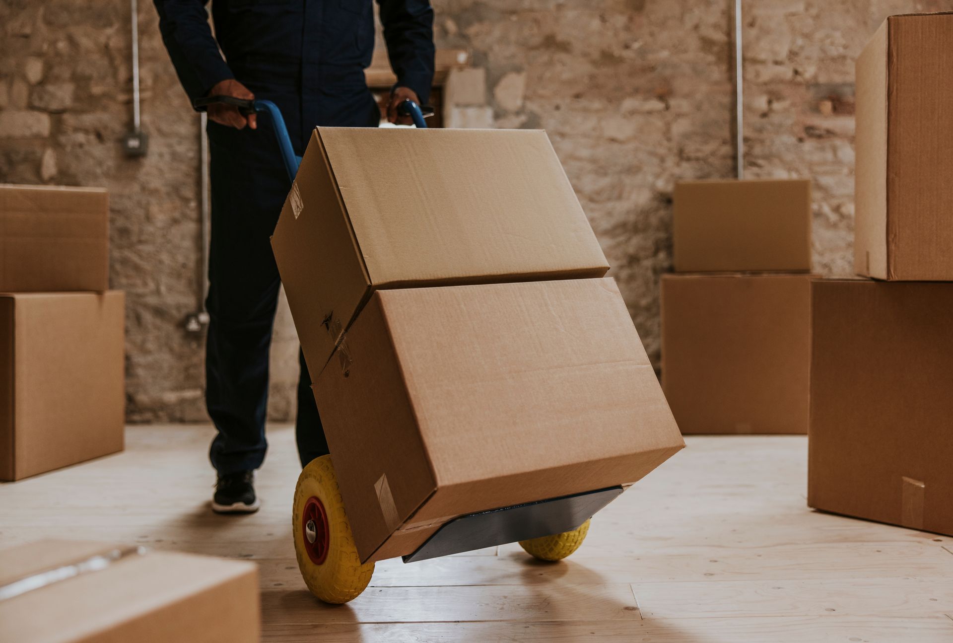 Person in a dark jumpsuit moving cardboard boxes with a hand truck inside a room with brick walls.