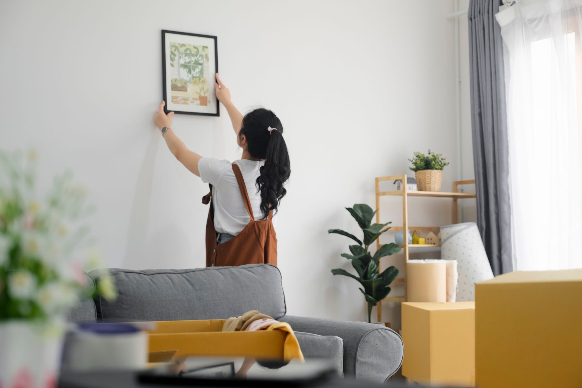 Woman hanging a framed picture on a white wall while unpacking moving boxes in her new living room.