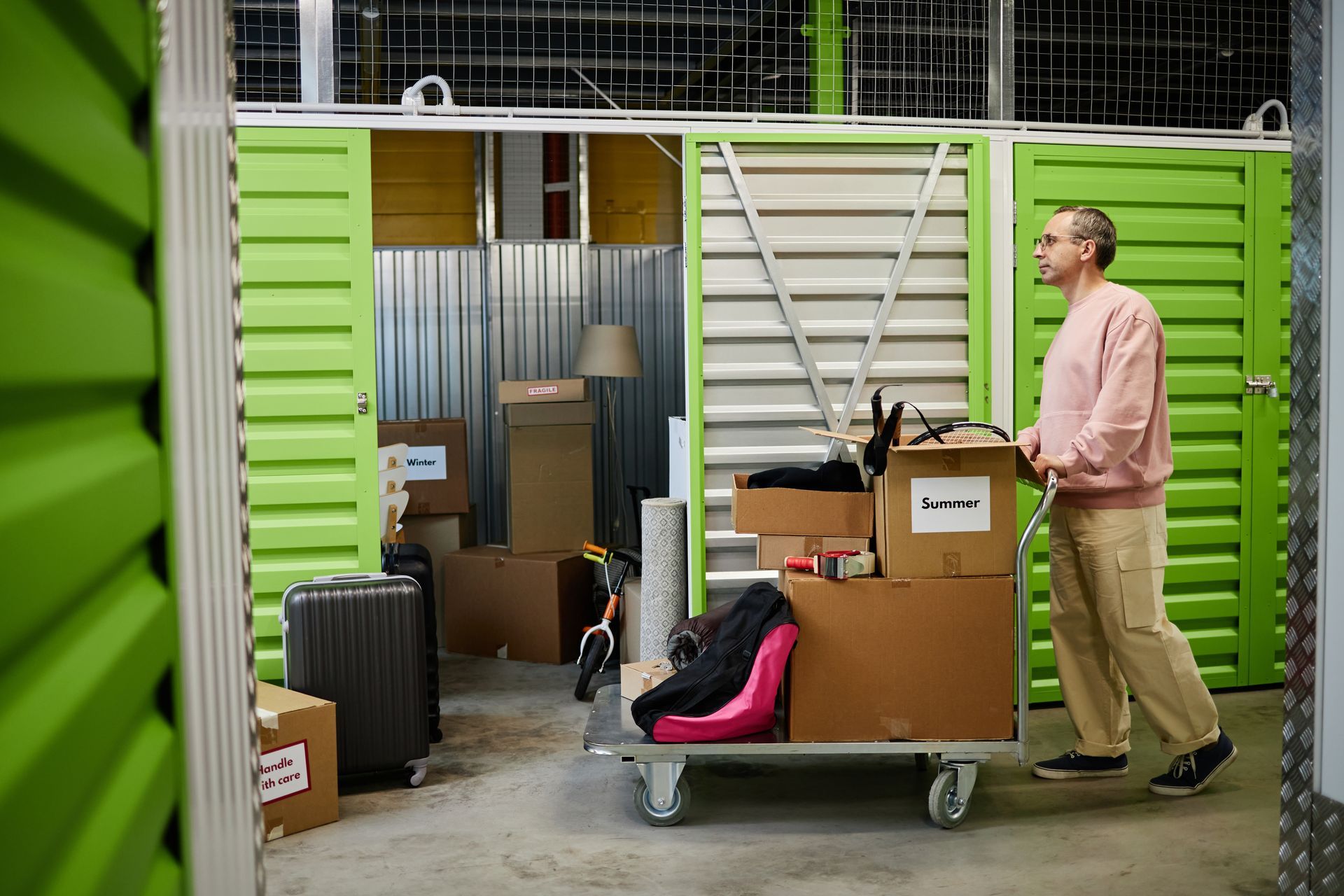 Man pushing a metal cart loaded with packed boxes and belongings into a green self-storage unit.