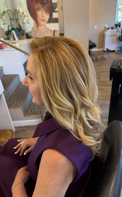 Woman with shoulder-length blonde hair seated in a salon, wearing a purple top, viewed in profile.