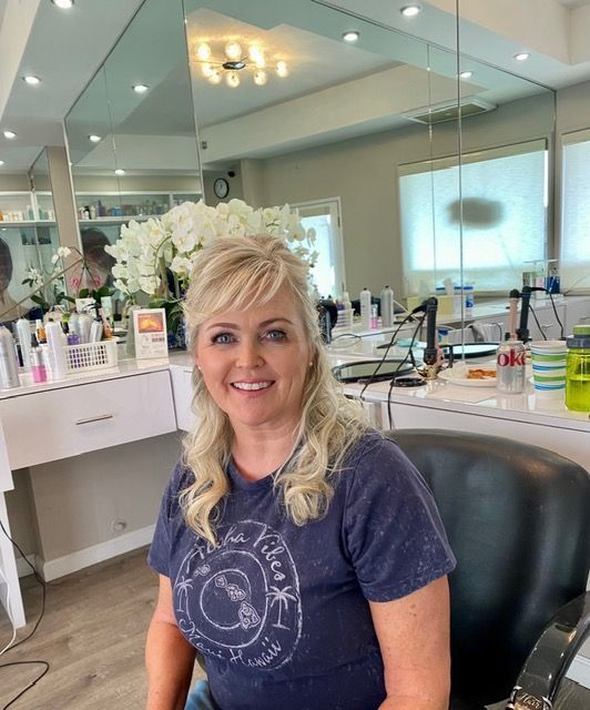 Smiling woman seated in a salon chair with styled blonde hair, in a bright mirrored hair salon.