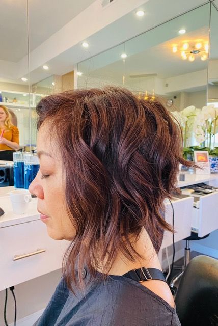 Profile view of a woman with short wavy auburn hair in a salon chair, eyes closed, with mirrors and lights behind her