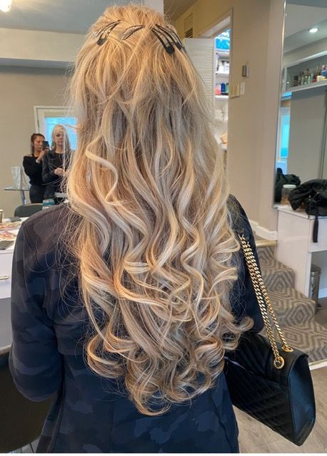 Back view of a person with long wavy blonde hair in a salon mirror area, holding a black handbag.
