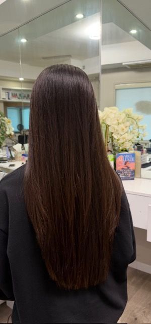 Woman seen from behind with long straight brown hair in a salon mirror, seated in a black cape