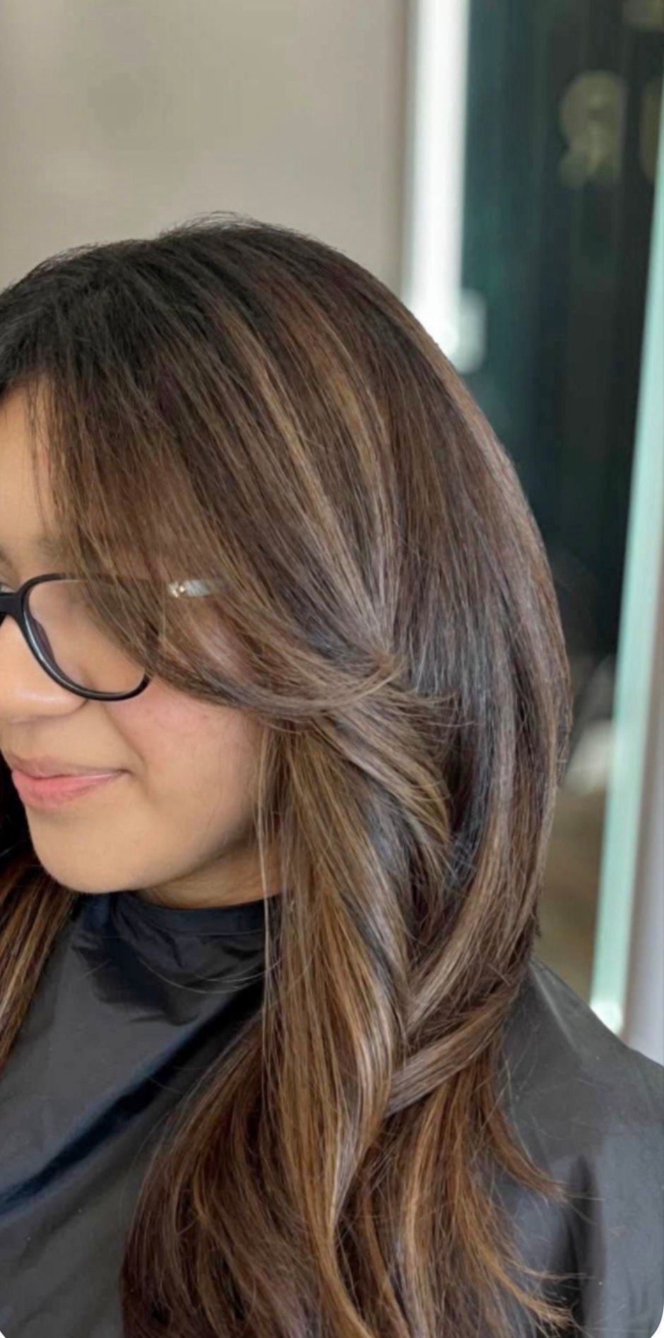 Woman in glasses with shoulder-length brown hair, wearing a black top, smiling in profile indoors.