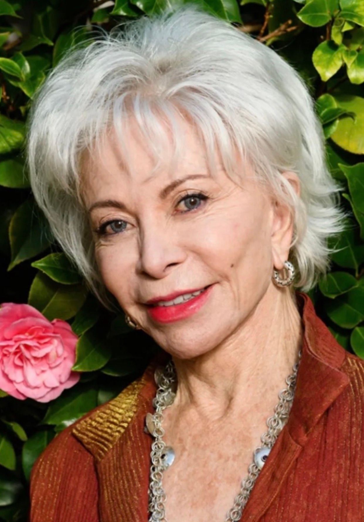 Portrait of a smiling woman with white hair beside a pink flower and green leaves