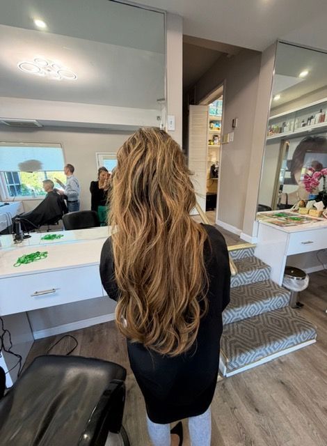 Person with long wavy hair stands at a salon vanity, seen from behind, with mirror and styling tools visible.